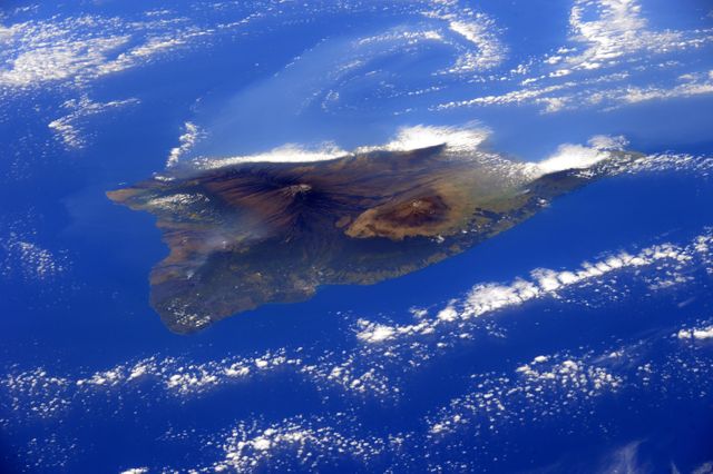 Stunning aerial view of Big Island Hawaii surrounded by the clear blue waters of the Pacific Ocean. Ideal for travel websites, educational materials about geography and nature, promotional content for vacation destinations, and nature-related publications. The cloud cover adds a dynamic and beautiful element to the scenery.
