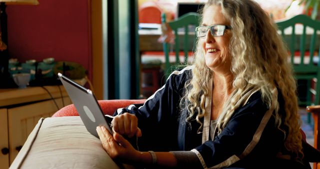 Senior woman using a tablet device while sitting on a couch in a well-lit living room. Ideal for themes related to senior lifestyle, technology adaptation, relaxed home environments, and older adults staying connected digitally.