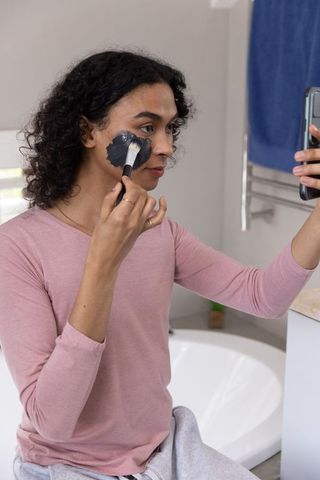 Nonbinary person applying charcoal face mask while checking phone on bathtub rim self care