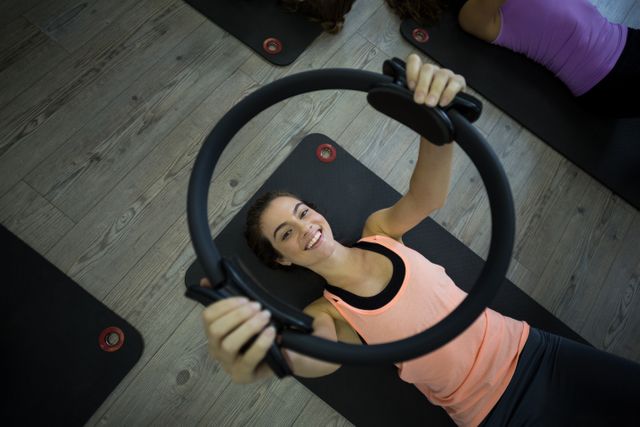 Fit woman lying on yoga mat, smiling while exercising with pilates ring in gym. Ideal for promoting fitness classes, healthy lifestyle, workout routines, and gym memberships.