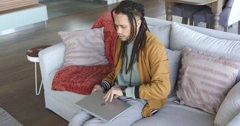 Man with dreadlocks using laptop on couch in cozy modern living room interior