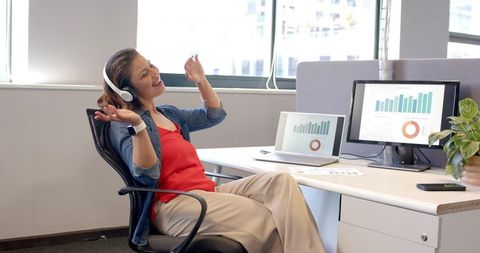 Professional woman celebrating at desk while listening to headphones with laptop and charts
