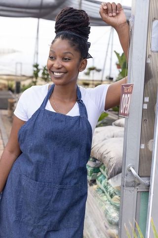 Smiling african american woman wearing denim apron in greenhouse doorway with open sign