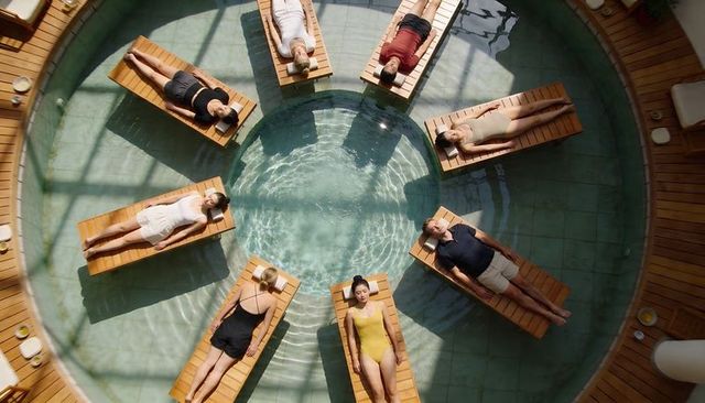 Eight adults relaxing on wooden loungers around circular pool under sunlit glass atrium