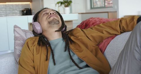 Young man with dreadlocks reclining on sofa wearing pink headphones listening to music