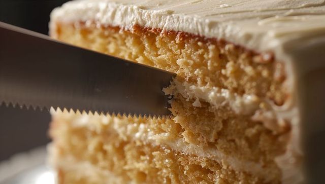 Slicing vanilla layered cake with serrated knife revealing moist crumb and cream filling
