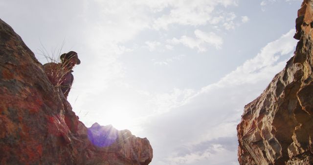 The image portrays an adventurer sitting on the edge of a rocky cliff, looking into a bright and clear sky. The person, wearing a hat, is positioned amidst rugged rocks with sunlight streaming through, suggesting a sense of solitude and connection with nature. This can be used for themes of exploration, nature, adventure, contemplation, or personal achievements. Ideal for outdoor adventure blogs, travel agencies, motivational posters, or environmental campaigns.