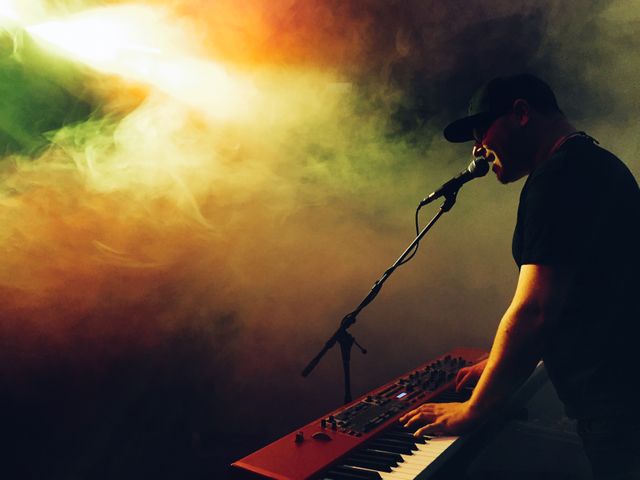 Male musician playing an electric keyboard and singing into a microphone on a dimly lit stage, surrounded by vibrant colored smoke effects. Perfect for music event promotions, concert posters, and entertainment-related content.