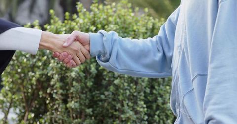 Multiracial colleagues shaking hands outdoors beside trimmed hedge, suit cuff and hoodie