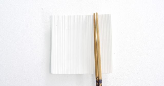 A pair of wooden chopsticks rests on a white, textured ceramic plate against a plain background, with copy space. The simplicity of the composition emphasizes the minimalistic aesthetic and the cultural utensil used in Asian cuisine.