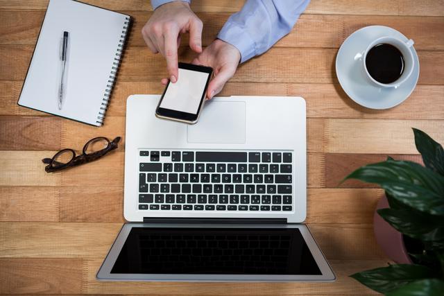 Businesswoman is using a mobile phone at her office desk, which features a laptop, notebook, pen, coffee, and plant. This can be used for themes related to professional work environments, office productivity, business communication, and corporate lifestyles.