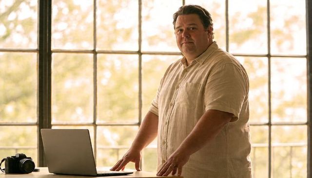 Confident entrepreneur standing window-side desk with laptop and dslr in warm golden light