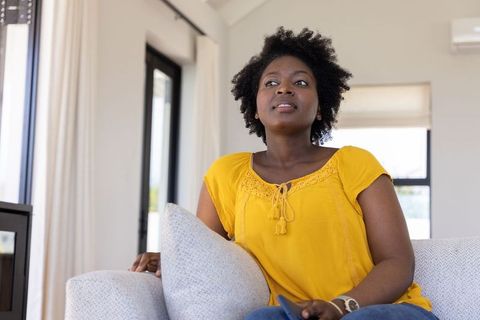 African american woman sitting on sofa in mustard blouse looking contemplative at home