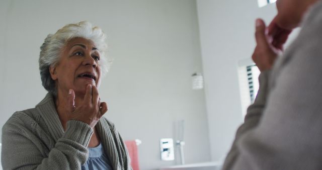Senior woman examining her face in a bathroom mirror, possibly checking for aging signs or skincare routine. Can be used in contexts related to elderly health, skincare products for seniors, daily routines, and aging concerns.