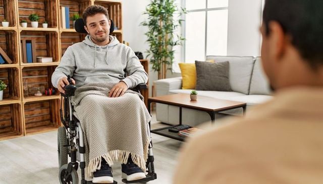 Young man navigating motorized wheelchair at home using joystick, relaxing under blanket