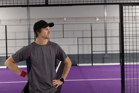 Male athlete standing confidently on indoor purple paddle tennis court wearing cap and wristwatch