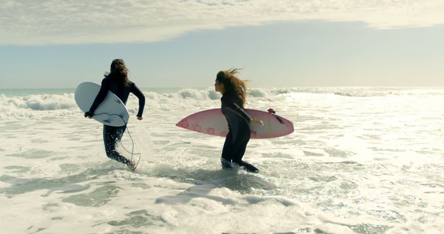 Two young Caucasian women are seen entering the ocean with surfboards, ready to enjoy a surfing session. Their enthusiasm and active lifestyle are captured as they prepare to ride the waves under the bright sun.