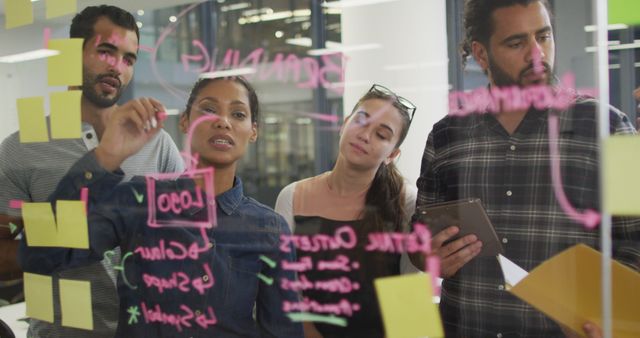 Team of diverse young professionals collaborating and brainstorming ideas using sticky notes on a glass wall in a modern office environment. Useful for ideas related to teamwork, business strategy, creative process, startup dynamics, and office interaction.