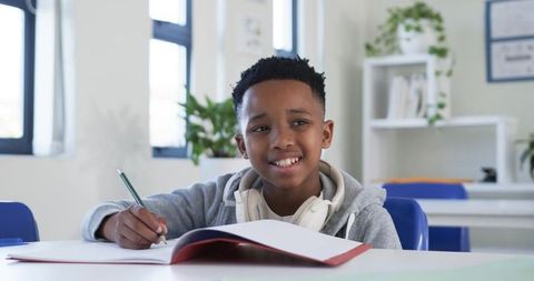 African american preteen student writing in notebook with headphones in bright classroom