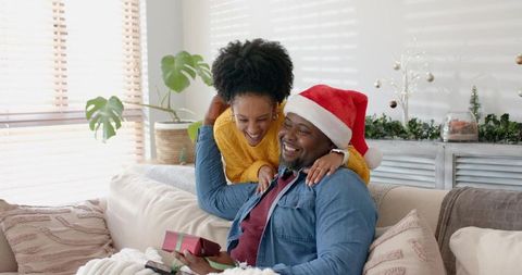 Diverse couple exchanging christmas gift on cozy living room sofa wearing santa hat
