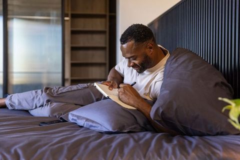 Mature african man writing in notebook while reclining on modern gray bed for journaling