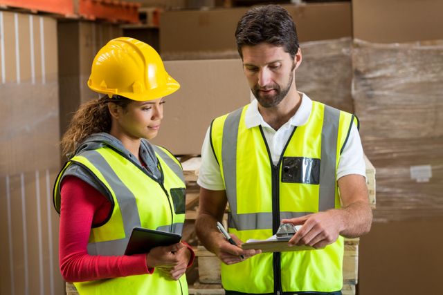 Warehouse workers in safety vests and hard hat discussing logistics using a clipboard and digital tablet. Ideal for illustrating teamwork, inventory management, supply chain processes, and industrial operations.