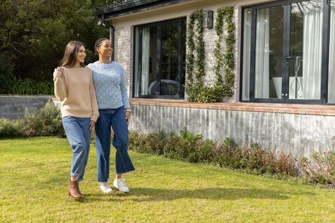 Diverse women walking arm-in-arm across sunlit lawn beside modern home with glass doors