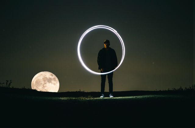 Person standing in open field at night, holding light source forming a ring shape. Full moon and dark sky creating a dramatic background. Perfect for creative art projects, conceptual themes, night photography, or cosmic-themed designs.