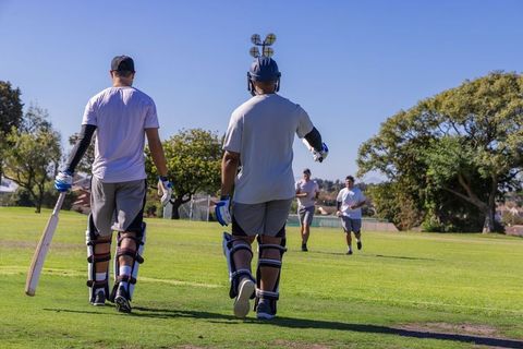 Two cricketers walking toward pitch on sunny green field with teammates running behind