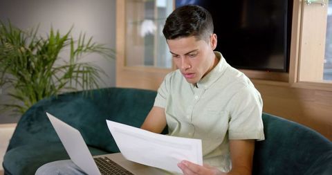 Young Man Reviewing Documents on Couch with Laptop in Home Office