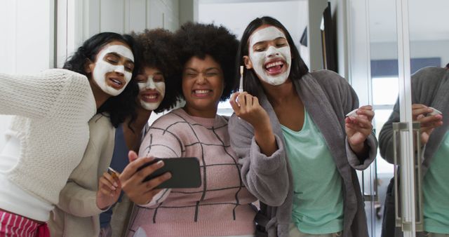 Four friends having fun during a spa day at home, wearing face masks and casual clothing, smiling and taking a selfie together. Perfect for themes of friendship, self-care, relaxation, and joyful moments. Can be used in blog posts about skincare tips, personal care routines, or advertisements for beauty products.