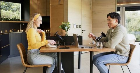 Young cohosts recording podcast at dining table in modern kitchen with microphones, laptop