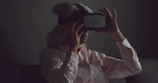 Elderly woman is experiencing virtual reality by using a VR headset in a dimly lit room. Ideal for highlighting senior engagement with modern technology, showcasing advancements in VR experiences suitable for all ages, or emphasizing tech accessibility for elderly population.