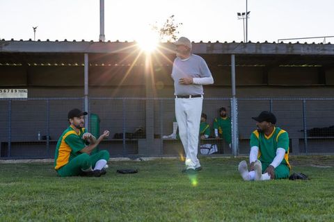 Senior coach guiding male cricket team at sunset on grassy field near dugout