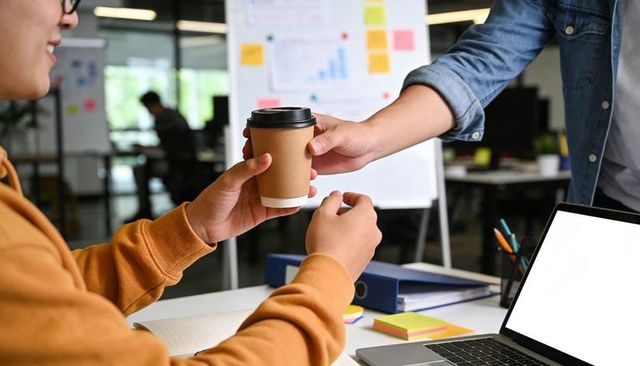 Coworkers passing coffee at open office desk showing casual collaboration and teamwork