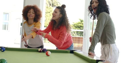 Multicultural women playing pool in sunlit home gameroom, cheering and pointing at shot