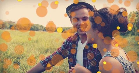 Laughing young couple leaning close in sunlit meadow wearing fedora and floral dress