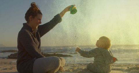 Mother pouring sand over toddler on sunset beach, backlit playful bonding moment