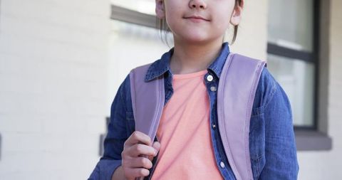 Confident schoolgirl holding backpack strap wearing denim jacket and tee urban portrait