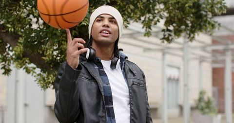 Young man spinning basketball on finger wearing beanie and headphones on campus walkway
