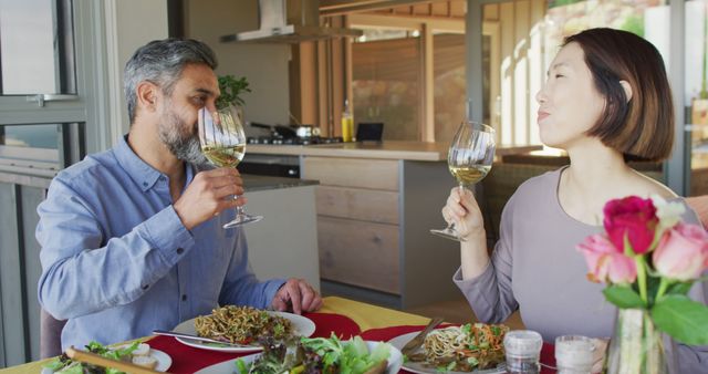 Couple enjoying dinner and wine together at a cozy dining table in a modern apartment. Both are engaged in conversation while savoring their meal which includes a salad and noodles. Perfect for concepts related to lifestyle, home dining, romance, and social interactions. Ideal for use in advertisements, websites, and publications focusing on relationships, domestic life, or dining experiences.