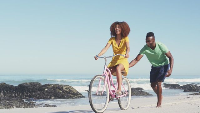 A joyful scene featuring an African American couple enjoying their vacation on a tropical beach. The woman is riding a bike, while the man is running behind her, pushing and laughing. This captures a moment of playful activity and happiness by the coastline. This vibrant image is perfect for use in advertisements or articles about vacation travel, outdoor adventures, beach activities, young couples, and healthy lifestyle inspiration.