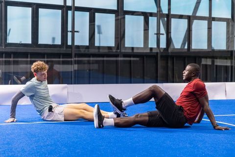 Male friends stretching hamstrings on blue turf in indoor sports facility