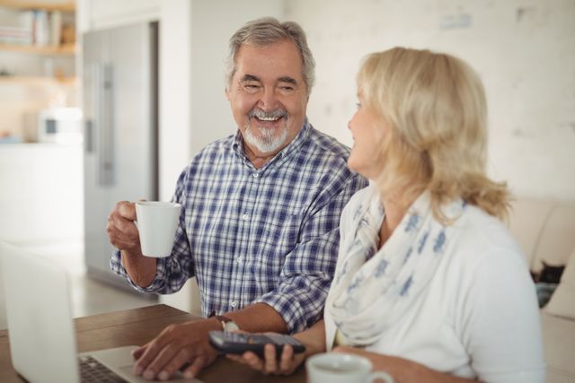 Senior couple sitting at home, enjoying coffee while using a laptop. Perfect for illustrating themes of retirement, technology use among seniors, and happy home life. Suitable for articles on senior lifestyle, technology adoption, and family bonding.