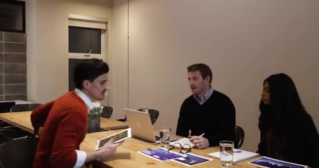 A young man in a red sweater sitting down with two colleagues for a business meeting in a modern office. They are discussing at a wooden table with documents, notebooks, and laptops on it. Ideal for content related to business teamwork, corporate meetings, professional collaboration, and office environments.