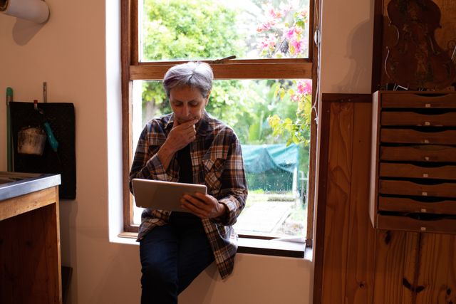 Front view of a senior Caucasian female luthier sitting on the windowsill, holding computer tablet in her workshop with tools hanging up on the wall in the background.