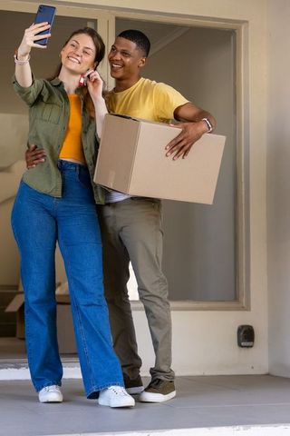 Young couple celebrating move-in taking selfie with keys and carrying moving box