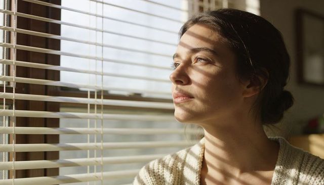 Young woman gazing through window blinds catching striped sunlight wearing knit sweater