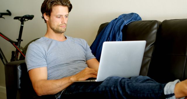 Man is sitting on a black leather sofa using a laptop while resting his legs on the sofa. He is wearing a grey t-shirt and jeans. Behind him, a bicycle is partially visible, suggesting an active lifestyle. The relaxed and casual home environment makes this suitable for concepts related to remote work, relaxation, technology at home, and lifestyle balance.