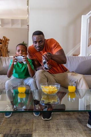 African American Father and Son Playing Video Games on Couch with Snacks and Juice